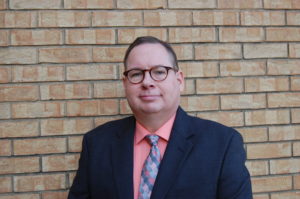 A man in glasses and a suit standing next to a brick wall.