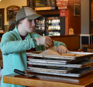 A woman in a hat is putting toppings on some pizza.
