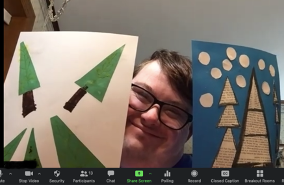 young man in glasses holds up art made in the Artist In You Class during a Zoom call.