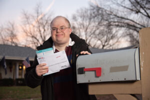 A man in a jacket, wearing glasses, stands in front of a home near his mailbox. He is smiling and holding mail.