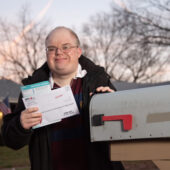 A man in a jacket, wearing glasses, stands in front of a home near his mailbox. He is smiling and holding mail.