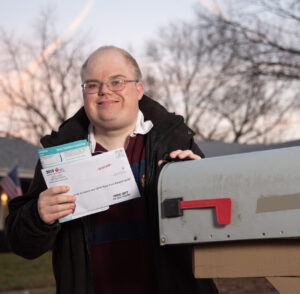 A man in a jacket, wearing glasses, stands in front of a home near his mailbox. He is smiling and holding mail.