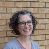 woman with glasses, smiling, against a brick wall wearing a purple shirt and grey cartigan