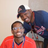 One young man with glasses sits wearing a red shirt, another man is standing but leans in close and smiles, he's wearing a cardinals hat and blue shirt
