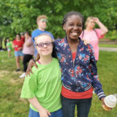 two young girls in the park, one taller girl with a blue jacket and red shirt has her arm around the other girl who is wearing glasses and a green shirt, other people are walking behind them in the distance through the park