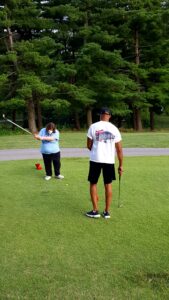 A woman, masked in a blue shirt, looks down with her golf club raised to hit the golf ball in front of her. A man, in a white shirt and black shorts, stands with his back to the camera and holds a golf club in his right hand.