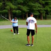 A woman, masked in a blue shirt, looks down with her golf club raised to hit the golf ball in front of her. A man, in a white shirt and black shorts, stands with his back to the camera and holds a golf club in his right hand.