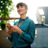 woman in teal shirt with a smile has her phone in her hand and the sun setting behind her
