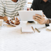 two people pictured from shoulder to waist, sit at a table while reviewing something on a tablet