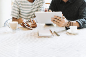 two people pictured from shoulder to waist, sit at a table while reviewing something on a tablet