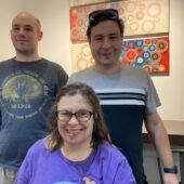 Three members smile in the front sits a woman with glasses, brown hair, and a purple shirt, in the back on the left is man with a long sleeve grey shirt, in the back right is a young man with brown hair, sunglasses on top of his head and a grey, white and black striped shirt.