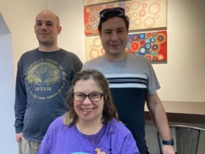 Three members smile in the front sits a woman with glasses, brown hair, and a purple shirt, in the back on the left is man with a long sleeve grey shirt, in the back right is a young man with brown hair, sunglasses on top of his head and a grey, white and black striped shirt.