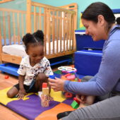 a toddler in a white shirt stacks blocks with a woman in a purple shirt and hair pulled back in a ponytail