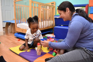 a toddler in a white shirt stacks blocks with a woman in a purple shirt and hair pulled back in a ponytail
