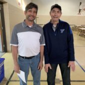 Two men stand and pose for a photo. One man with brown hair, a mustache and beard is wearing a white and grey polo, the other man is clean shaved wearing a hat and a navy pullover.