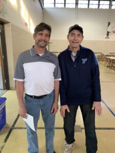 Two men stand and pose for a photo. One man with brown hair, a mustache and beard is wearing a white and grey polo, the other man is clean shaved wearing a hat and a navy pullover.