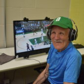 An older man sits in front of a computer with headphones on watching a basketball game.
