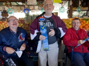 Three older men pose in front of a farmers market