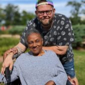 An older woman sits in a wheelchair with a man standing behind her. They hold hands over her shoulder and smile.