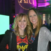 two women pose for a photo in front of neon lights and the Arc logo projected on the wall.