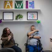 A woman and a man sit while stretching their arms, above them are three canvas paintings spelling AWE