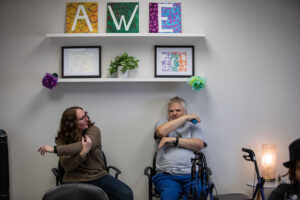 A woman and a man sit while stretching their arms, above them are three canvas paintings spelling AWE