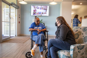 A woman sits in a wheelchair speaking with another woman sitting on a couch wearing a mask.