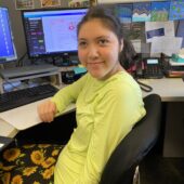 A young woman smiles for the camera while she sits at a computer doing research